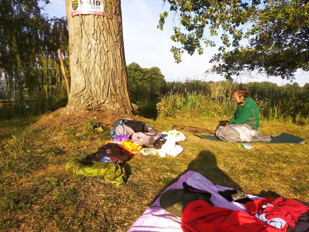 Rozrzucone ubrania i inne elementy biwaku na trawie. W lewym górnym rogu drzewo z tabliczką ochorny.