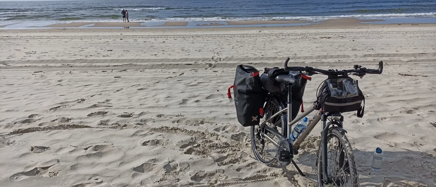 Zdjęcie przedstawia rower z sakwami na pustej plaży. W tle Morze Bałtyckie.