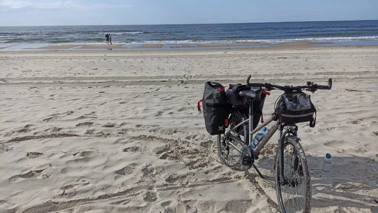 Zdjęcie przedstawia rower z sakwami na pustej plaży. W tle Morze Bałtyckie.